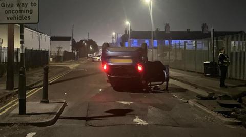 A blue car on its roof, with an open door and red rear lights shining, in a narrow road in central Hull. In the foreground the road narrows around a small speed bump. A police officer stands to the right and a police car can be seen in the background. 