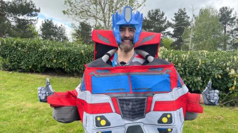 A man with a beard is smiling at the camera with his thumbs up. He is wearing an outfit for the character Optimus prime 