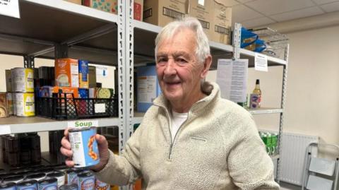 A man with grey hair and a white jumper holding a cup of beans in one hand and standing in front of shelves holding more tins