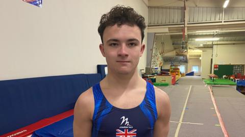 A young man standing inside a gymnasium wearing a UK navy blue leotard looking directly at the camera. Tumbling mats and other gymnastic equipment is visible behind him.