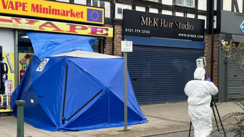 A blue tent covers the scene outside a vape shop. A scenes of crime officer in a white protective suit is in the foreground.
