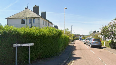 A residential street with cars parked and a hedge outside the houses. A street sign saying Dronley Avenue is in the foreground