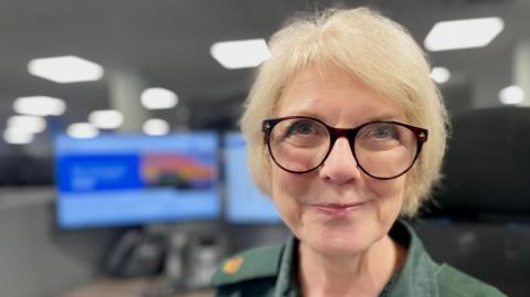 A woman in ambulance service uniform wearing glasses sitting in front of a 999 call handlers computer screen