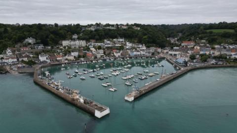 An ariel view of St Aubin in Jersey at high tide. There are boats in harbour and there are house surrounding the harbour. In between the buildings is green trees and green hills in the distance. 