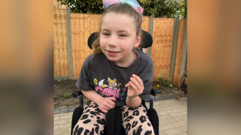 Ada wearing leopard print style leggings and siting in her wheelchair. She is leaning forward, smiling at the camera, and wearing a grey t shirt with an animation cat character on. Behind her is a garden fence.