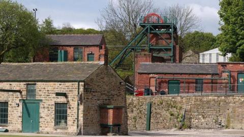 A general view of buildings at the National Coal Mining Museum in Wakefield. The building in the foreground has beige coloured bricks with a green door. Several red brick buildings stand in the background, with green doors and railings, with the Museum's red pit wheel at the top of the tallest building.