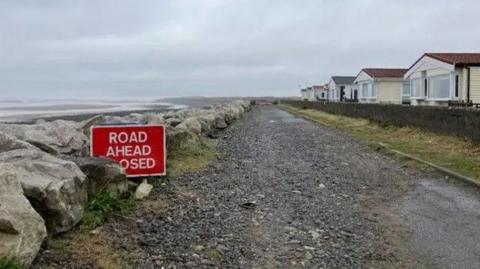 A dirt road with large rocks lining the left side with the sea behind them. Static caravans can be seen to the right of the road.