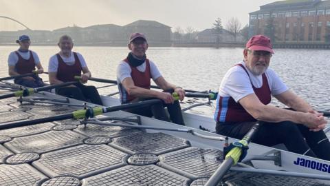Four men with white, red and blue tops, three with caps, sitting in a rowing boat on the water. Buildings can be seen behind them.