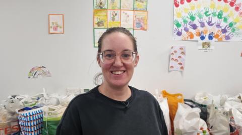 Carlisle foodbank's operations director Steph Humes inside the foodbank. She has brown hair tied back and wears glasses. She is wearing a black t-shirt and is smiling at the camera. Behind her are supermarket-branded bags filled with food. On the wall there are children's drawings and paintings.