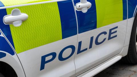 The side of a police car with the words POLICE in capital letters written on the side of it in blue. The traditional police markings are also on the car.