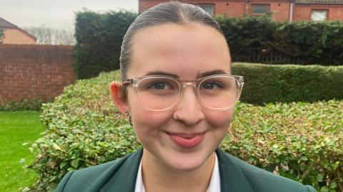 A schoolgirl who has long brown hair tied back and is wearing a green school uniform, clear-framed glasses as she stands in a garden.