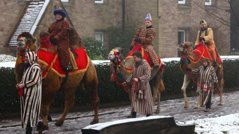 Three camels with red cloths covering their backs and colourful bridles, being ridden by three people dressed as wise men, led by two men and a woman in striped robes. They are walking on a snowy path past brick dwellings.