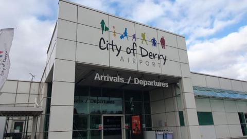 Front of City of Derry Airport building. It has graphics of people above the City of Derry Airport sign on a white building. There is an Arrivals/Departures sign above a glass entrance door.