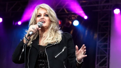 Bonnie Tyler singing on stage, wearing a black jacket and holding a microphone in her right hand