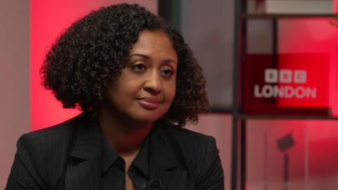 Andrea has short, dark curly hair and wears a dark, pinstripe suit. She is sitting in front of a red, BBC London branded backdrop.