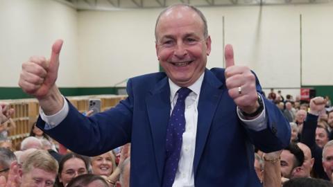 Fianna Fail leader Micheál Martin is hoisted up by supporters. He smiles and gives the thumbs up. He is wearing a blue suit with a white shirt and blue tie.