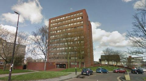 Warwick Court is a large 11-storey building made out of brown brick. Rows of windows line each floor. Several trees stand outside the building.