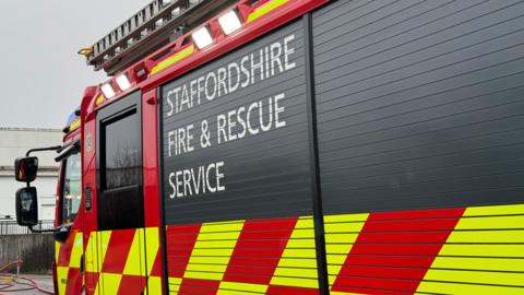 A side view of a fire engine. There is white lettering on a black roll-down panel which says Staffordshire Fire & Rescue Service.