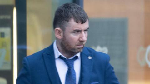 A man is seen in a navy jacket and blue tie at a court appearance. 
He has short brown hair and stubble.  