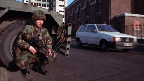 An army soldier is crouched down wearing a uniform and holding a rifle as he lays against an armoured vehicle. A car drives past him.