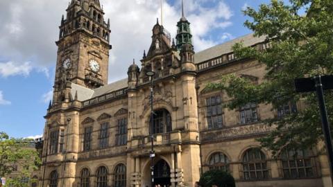 Sheffield Town Hall. It is an old-fashioned, stone build building. It has a clock tower and some turrets on the corners of the building. There is a balcony above the main entrance.