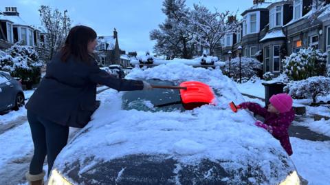 A mother and daughter clear the snow-laden windscreen of a car in the early hours. The car is on an upmarket residential street with sandstone properties, trees and bushes