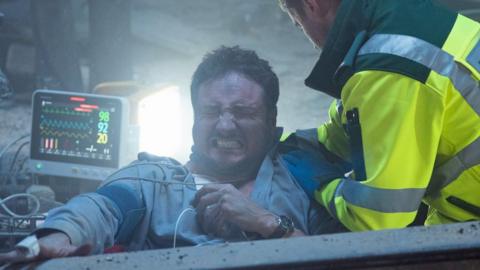 Martin Fowler (James Bye) wincing in agony helped by a paramedic in a fluorescent jacket