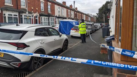 A policeman stands guard on a pavement, with blue and white tape cordoning off the area. A blue forensic tent stands in the middle of the street. Wood Green Road is a red-brick Victorian terrace with bay windows fronting on to the pavement and cars parked along the street