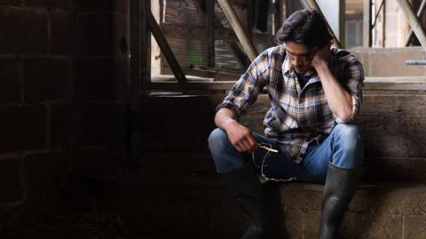 Stock photo of a young farmer sitting with his head in his hands in a barn