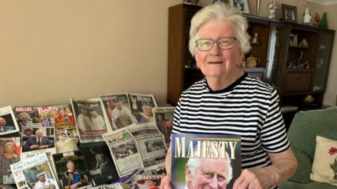 A woman holding a magazine featuring King Charles III on the cover. On the table behind her are various newspaper clippings featuring the Royal Family