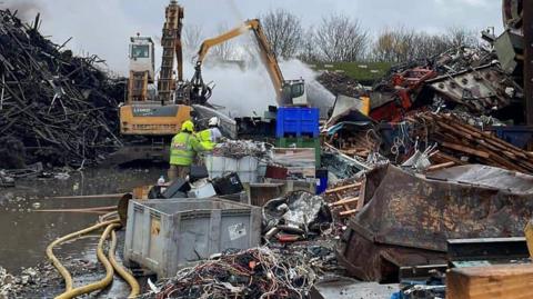A crane is moving metal and yellow hoses are in the picture. There are also large piles of scrap metal and smoke can also be seen. Near the camera are large items of scrap metal, including boxes, sheets and blocks of metal. 