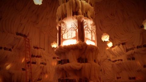 A warmly lit, intricate sculpture resembling a lantern amidst walls adorned with fluffy white formations and glowing accents.