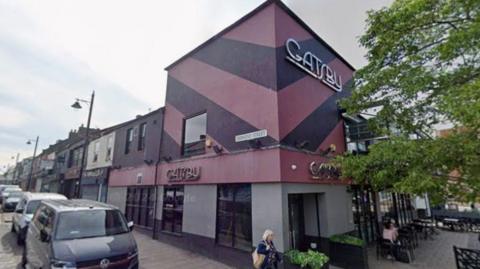 Streetview of Gatsby, a bar in Sunderland. It is a two storey block of a building painted with chevrons in two shades of purple, with trees and seats to the side of the picture. There are cars parked down the street and a woman with blonde hair is walking past. The street lamps are ornate.