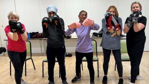 A group of women and a man with a turban hold their arms up whilst wearing boxing gloves. Various equipment and chairs can be seen in the background behind them.