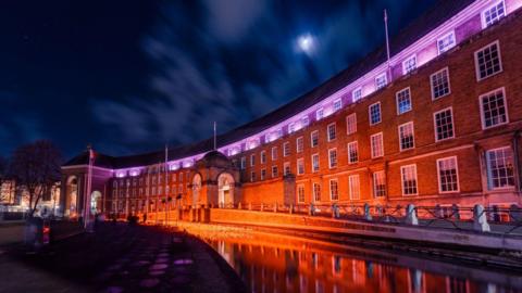 Bristol City Council House seen at night. The front of the building is illuminated by orange and pink lights. The Council House is reflected in the water in front of the building.