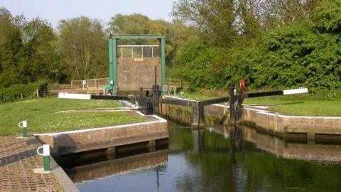 A canal lock with gates and a metal lift bridge surrounded by greenery on a sunny day.