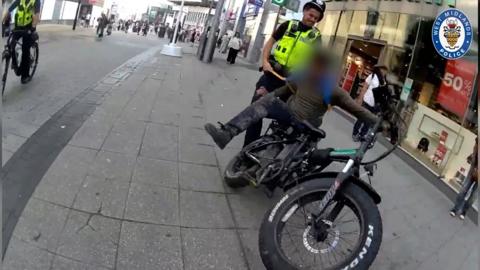 A black e-bike is stopped on a pavement with a police officer standing behind it.