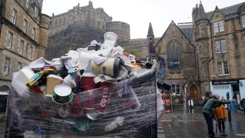 An overflowing bin in Edinburgh's Grassmarket. Several coffee cups and food containers are wrapped around the top of the black bin with cellophane. The sky is overcast and in the background, Edinburgh Castle can be seen on the hill.