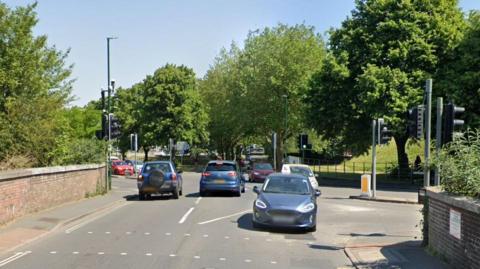 Cars driving on the four-way junction at Maumbury Cross in Dorchester. To the left is a brick wall with a narrow pavement.