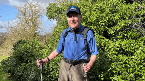Mike Patrick on a sunny day outdoors in nature with greenery in the background. He is wearing a blue polo shirt, a Cure EB cap and is carrying walking poles. 