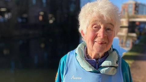 Barbara Thackray, with white hair, smiles as she is photographed in a light blue running vest on top of a purple fleece and grey coat. She is standing by the side of a canal on a sunny day.