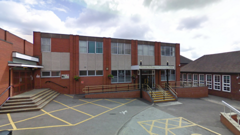 The exterior of a red-brick school building with steps and ramps leading up to the doors. There are yellow lines marked out on the car park in front of the building.