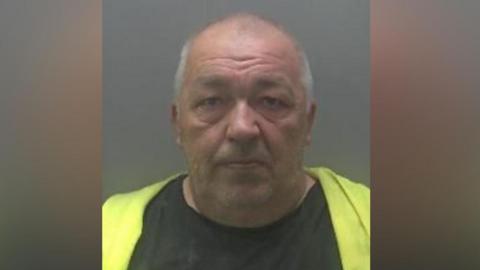 Custody image of Mindaugas Borisas, a man, with short white hair, wearing a black T-shirt and a green jacket.