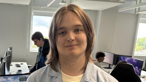 Alfie Johnson is standing in a room where there are several computers. Johnson has shoulder length brown hair and is wearing a yellow T-shirt and blue shirt.