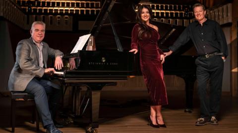 Lee Michael Walton, a man with short grey hair wearing a grey suit jacket and dark trousers, sits at a black grand piano with a metronome on it. Monica McGhee, a woman with long dark hair wearing a dark red velvet dress, stands to the right of the piano with her hand on it. Toby Spence, a man wearing a dark shirt and dark trousers, stands to on the far right with his hand on the piano