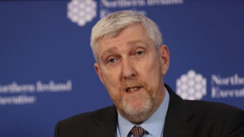John O'Dowd is wearing a blue shirt with a red and green tie. He has a black blazer on. He has grey hair and grey facial hair. Behind him is a blurred blue background with white logos.