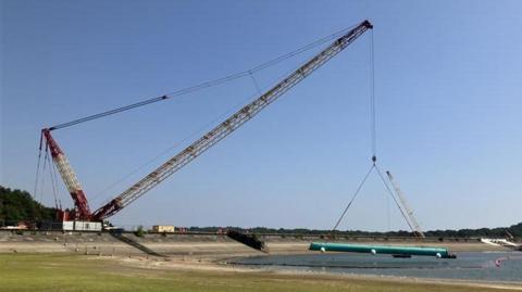 A large crane lifting a large blue pipe above a pool of water. The crane is white and red and is set against a clear blue sky.