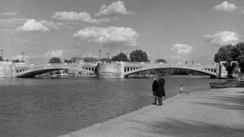 A black and white picture of Caversham Bridge and the River Thames, with two boys stood on the edge of the river.