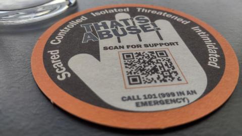A round beer mat on a grey table. It is coloured black, white and orange and features a white illustration of a hand, along with a QR code and the messages: "That's Abuse!"; "Scared, controlled, isolated, threatened, intimidated"; "Scan for support"; and "Call 101 (999 in an emergency)". 