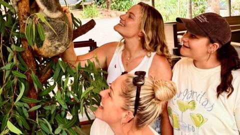 Three women looking at a Koala sat in a tree. The three women are all smiling.